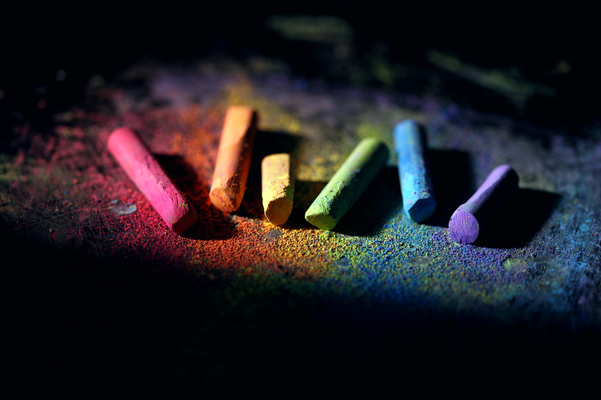 Photo of six pieces of chalk arranged in a rainbow against a dark background.